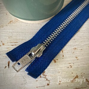 May include: A close-up of a blue zipper with a silver metal slider. The zipper is lying on a white wooden surface.