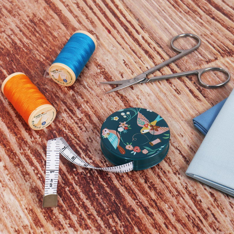 Puede incluir: Un kit de costura con carretes de hilo azul y naranja, tijeras peque&ntilde;as, una cinta m&eacute;trica y un recipiente redondo color turquesa con un dise&ntilde;o de p&aacute;jaros y flores. Sobre una superficie de madera.