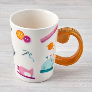 May include: White ceramic mug with a colourful sewing themed design. The mug has a yellow handle shaped like a measuring tape.