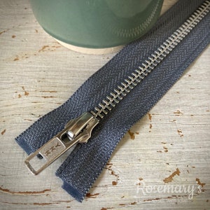 May include: A close-up of a dark gray zipper with a silver metal slider. The zipper is lying on a light wood surface.
