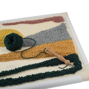 May include: A close-up of a rug hooking project in progress. The rug is made of a variety of colors, including yellow, orange, green, and gray. A wooden rug hooking tool is used to pull the yarn through the fabric. A ball of green yarn is also visible in the image.