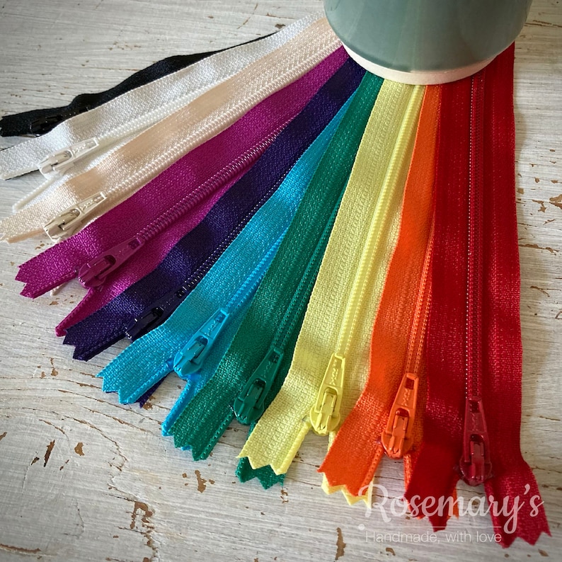 May include: A collection of 12 different colored zippers, arranged in a fan shape. The zippers are all closed and have a metal slider. The colors are black, white, cream, purple, dark blue, light blue, green, yellow, orange, and red.