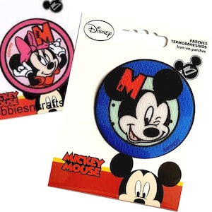 May include: Iron-on patch featuring Mickey Mouse with a red "M" on his ear. The patch is blue with a white background and has a white outline. The text "Mickey Mouse" is printed on the packaging.
