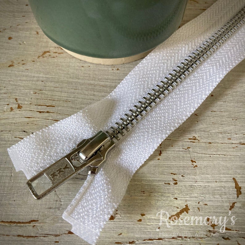 May include: A white zipper with a silver metal slider. The zipper is lying on a white wooden surface.