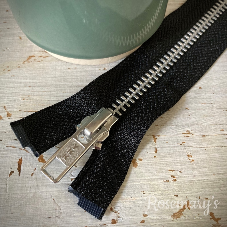 May include: A black zipper with a silver metal slider and teeth. The zipper is open and the slider is pulled down. The zipper is on a white wooden surface.