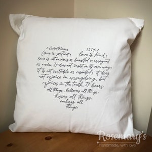 May include: A white square cushion featuring a heart-shaped arrangement of black script text, a quote from 1 Corinthians 13:4-7 about love. The cushion rests on a wooden surface and has the text "Rosemary's Handmade, with love".