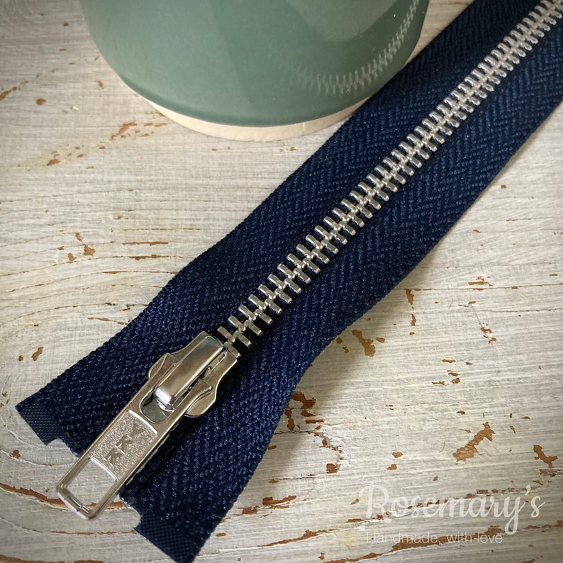 May include: A navy blue zipper with a silver metal slider and teeth. The zipper is lying on a white wooden surface.
