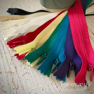 May include: Assortment of zipper tapes in various colors, including black, white, yellow, green, teal, purple, and red. The zippers are arranged in a fan shape, with the black zipper at the top and the red zipper at the bottom. The zippers are all closed and have a herringbone pattern.
