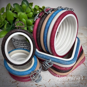 May include: A collection of circular and square embroidery hoops featuring textured outer frames in shades of dark brown, tan, blue, white, and red, paired with smooth white inner rings and metallic tension screws at the top of each frame.