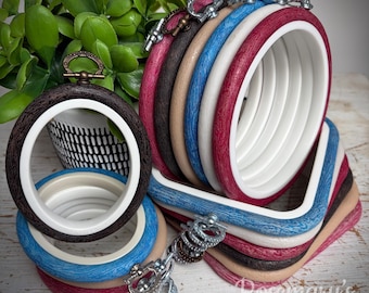 Puede incluir: Una colección de bastidores de bordado circulares y cuadrados con marcos exteriores texturizados en tonos marrón oscuro, beige, azul, blanco y rojo, junto con anillos interiores blancos lisos y tornillos de tensión metálicos.