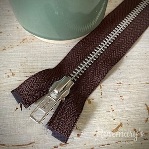 May include: A brown zipper with a silver metal slider. The zipper is lying on a white wooden surface.