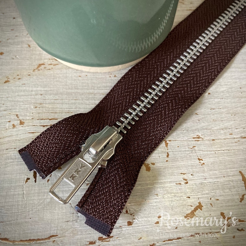 May include: A brown zipper with a silver metal slider. The zipper is lying on a white wooden surface.