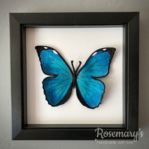 May include: Framed embroidered butterfly artwork. The butterfly has bright blue wings with black borders and white details. It's set against a white backdrop within a black frame. The text "Rosemary's Handmade, with love" is in the bottom right corner.
