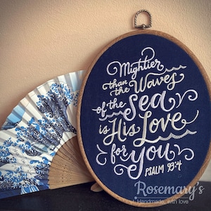 May include: Embroidered hoop art with a navy blue background and white embroidery. The text reads "Mightier than the waves of the sea is His Love for you."  Psalm 93:4. The hoop is surrounded by a wooden frame.