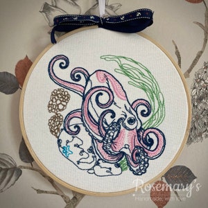 May include: An embroidery hoop containing white fabric with a stitched octopus design in pink and dark blue. The octopus is surrounded by green seaweed, brown coral, and a small blue starfish. A dark blue ribbon with white anchors hangs from the top.