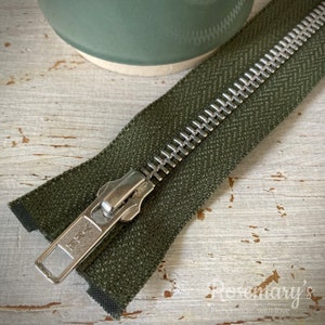 May include: A close-up of a green zipper with a silver metal slider. The zipper is lying on a light wood surface.