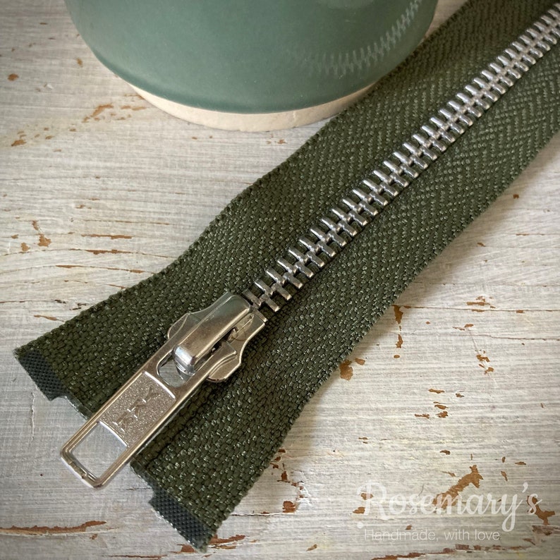 May include: A close-up of a green zipper with a silver metal slider. The zipper is lying on a light wood surface.