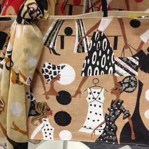 May include: A beige tote bag with a black and white patterned design featuring women in dresses and a scarf with white polka dots. The bag has a brown canvas texture and a black handle.