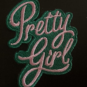 May include: An embroidered fabric patch featuring the text Pretty Girl in light pink cursive script lettering. The letters are outlined and set against a dark green background with a matching green stitched border edge.