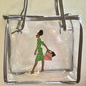 May include: Clear plastic tote bag with gray trim and a black and green embroidered design of a woman carrying a pink shopping bag with the text "1906" on it.