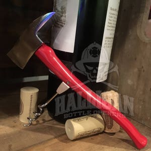 May include: A red and silver bottle opener shaped like a fire axe with a corkscrew attached. The opener is lying on a wooden surface with two wine corks.