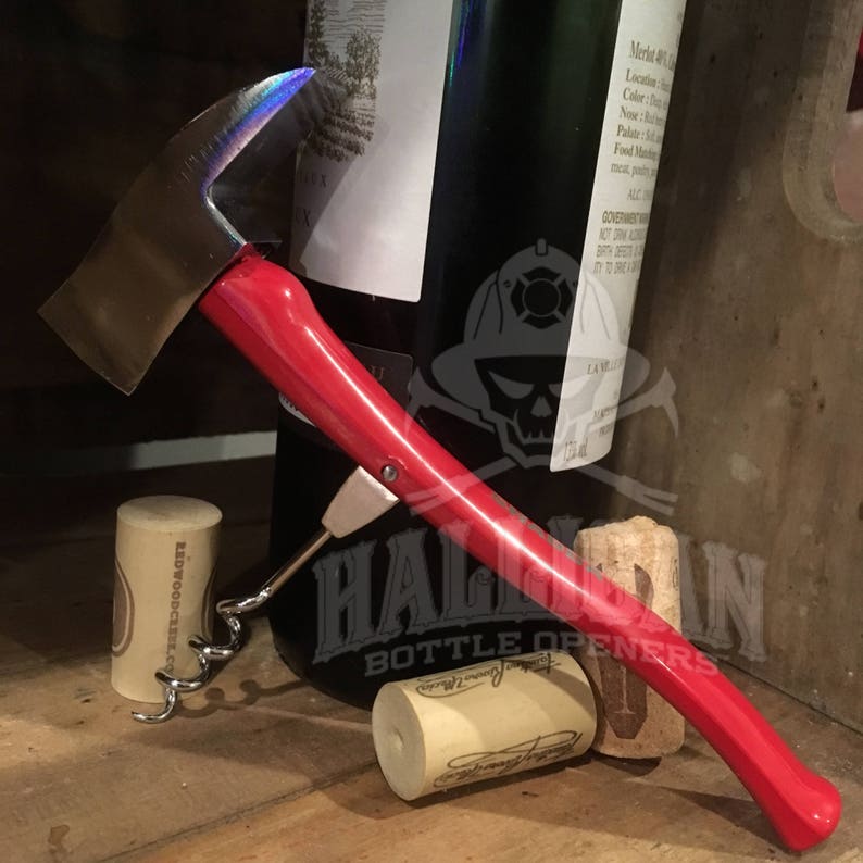 May include: A red and silver bottle opener shaped like a fire axe with a corkscrew attached. The opener is lying on a wooden surface with two wine corks.
