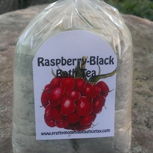 May include: A clear plastic bag filled with a gray bath tea blend. The bag has a label that says "Raspberry-Black Bath Tea" and features a drawing of a raspberry. The label also includes the website address "www.prettymommabeauty.etsy.com".