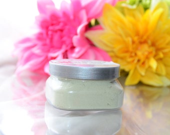 May include: A glass jar with a silver lid filled with a light green powder. The jar is sitting on a white surface with pink and yellow flowers in the background.