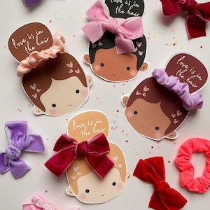 Puede incluir: Cuatro tarjetas ilustradas con caras de dibujos animados y el texto "love is in the hair". Cada tarjeta presenta un accesorio para el cabello diferente: un lazo de terciopelo rosa, un scrunchie morado, un lazo de terciopelo rojo y un scrunchie rosa.
