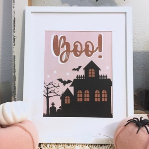 May include: A white framed print with a pink background and the word "Boo!" in brown script. A black silhouette of a haunted house with bats flying around it is in the center of the print.
