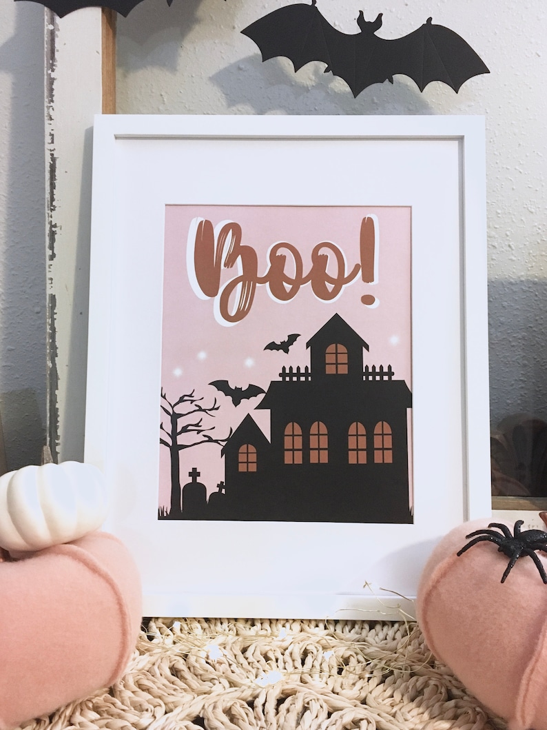 May include: A white framed print with a pink background and the word "Boo!" in brown script. A black silhouette of a haunted house with bats flying around it is in the center of the print.
