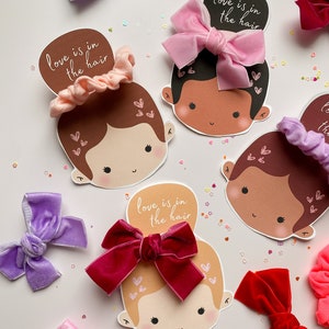 Puede incluir: Cuatro caras ilustradas con diferentes accesorios para el cabello, incluyendo una scrunchie rosa, un lazo de terciopelo rosa, un lazo de terciopelo burdeos y un lazo de terciopelo morado. Cada cara tiene el texto "love is in the hair" escrito encima.
