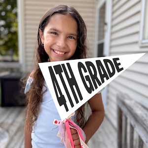May include: A white triangular pennant flag featuring the black text 4TH GRADE. The pennant is attached to a stick and decorated with several thin fabric ribbons in shades of pink, white, and patterned designs tied at the base of the flag.