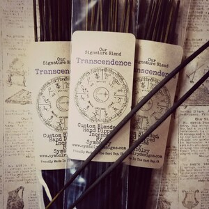 Pode incluir: Três pacotes de varetas de incenso pretas com etiquetas que dizem "Our Signature Blend Transcendence Custom Blended, Hand Dipped Incense By Symbolry www.symbolrydesigns.com Proudly Made In The East Bay, CA".