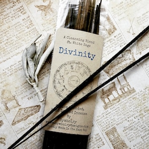 May include: A package of Divinity incense sticks with a white sage blend. The packaging features a brown label with the text "A Cleansing Blend ft. White Sage Divinity" and a circular design with astrological symbols. The incense sticks are bundled together and are ready to be burned.