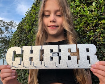 May include: Silver glitter "CHEER" sign with hooks for medals. The sign is held up in front of a green bush. The sign is silver with glitter and has the word "CHEER" in large letters.