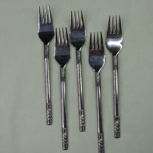 May include: Five silver forks with a decorative pattern on the handles. The forks have four tines each and are arranged on a light green surface. The handles have a detailed, woven-like design.