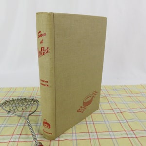 May include: A vintage cookbook titled "Serve it Buffet" with red text on a tan cover. The book has a textured appearance and features illustrations of plates. A silver-colored kitchen tool is in the foreground.