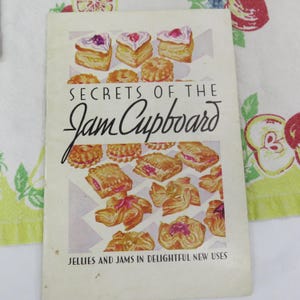 May include: A vintage cookbook titled "Secrets of the Jam Cupboard" with illustrations of pastries. The cover features various baked goods with jam fillings, including heart-shaped pastries. The text below reads "Jellies and Jams in Delightful New Uses."