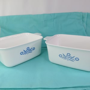 May include: Two rectangular white baking dishes with integrated side handles. Each dish features a blue floral pattern on the front, consisting of a central flower surrounded by leaves and smaller blossoms, centered on the smooth exterior surface.