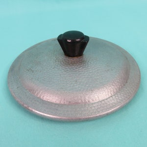 May include: A silver-colored, hammered metal lid with a black, art deco style handle. The lid is round with a slightly raised edge. The handle is a dark, matte black. The lid appears to be for a pot or pan.