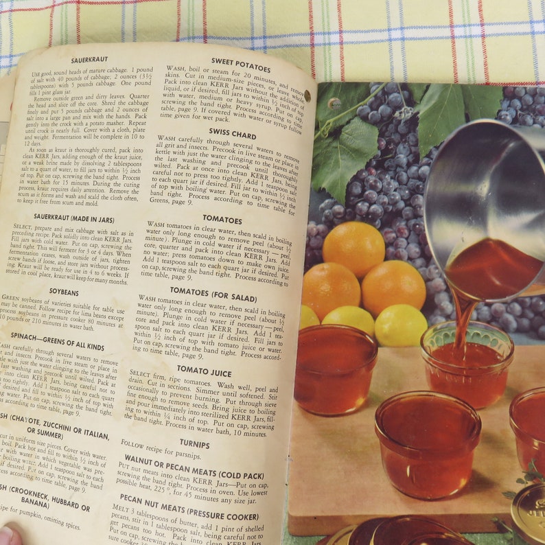 May include: An open cookbook displays recipes for canning tomatoes and other vegetables. Jars are filled with a red liquid, and oranges and lemons are nearby.