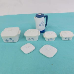 Puede incluir: Un conjunto de utensilios de cocina blancos y azules, que incluye varios recipientes cuadrados para hornear con tapas, una tetera y dos tapas. Los recipientes y la tetera presentan un diseño floral azul. Los artículos están dispuestos sobre una superficie azul claro.