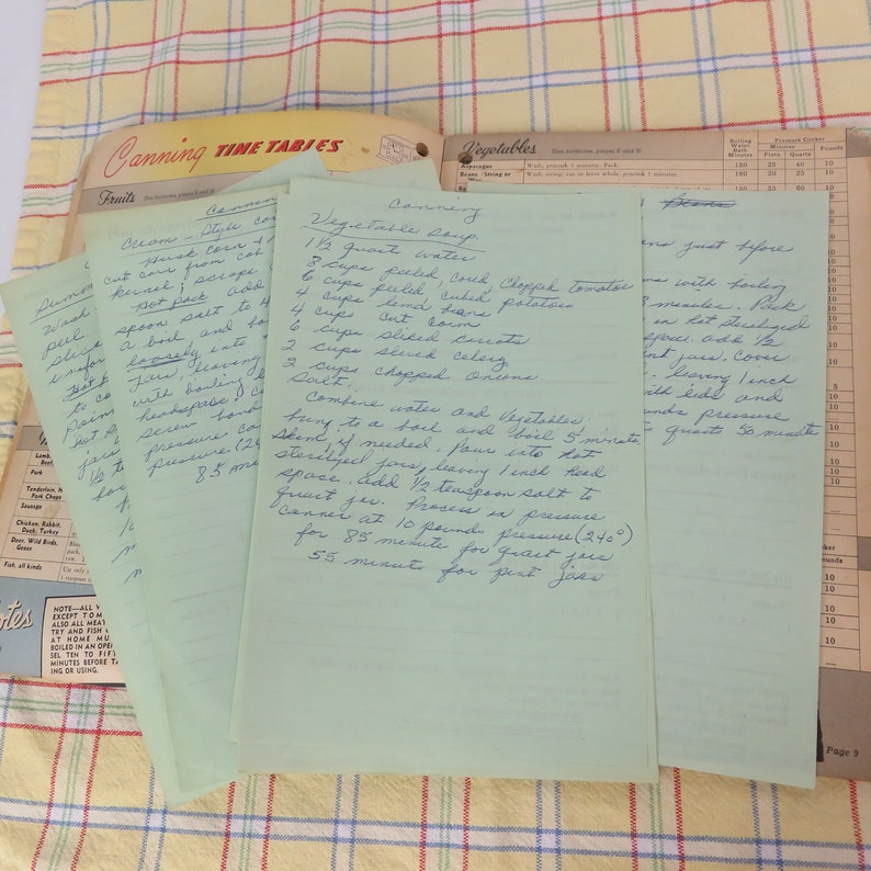 May include: A collection of handwritten recipes on light green paper, featuring canning instructions for vegetables. The recipes are on a yellow and white checkered tablecloth, with a vintage canning guide visible in the background.