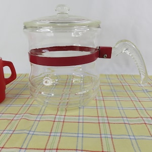 May include: A clear glass coffee pot with a lid and a red metal band around the middle. The handle is clear glass. The pot is sitting on a yellow and white plaid tablecloth.