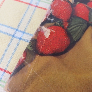 May include: A close-up of a vintage print featuring ripe red strawberries with green leaves. The print is torn and aged, with a brown background and a yellow and blue checkered tablecloth in the background.