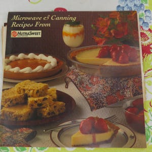 May include: A cookbook titled "Microwave & Canning Recipes From NutraSweet" is open, displaying images of various desserts. A pumpkin pie with whipped cream, a key lime pie with strawberries, and a plate of brownies are visible.