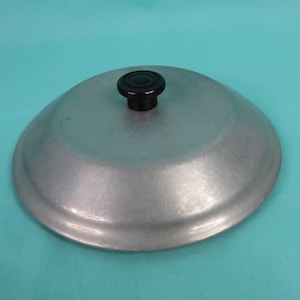 Puede incluir: Una tapa de olla vintage de aluminio con un pomo negro. La tapa es de color plateado claro y tiene una forma ligeramente abovedada. El pomo es redondo y de plástico negro. La tapa probablemente sea de un juego de utensilios de cocina.