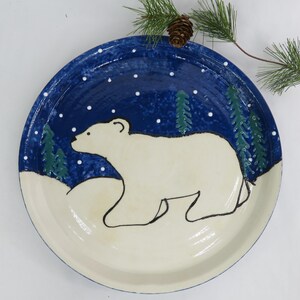 May include: A hand-painted ceramic plate featuring a polar bear in a snowy landscape. The plate has a dark blue background with white dots representing snowflakes and green trees. The polar bear is white with black outlines.