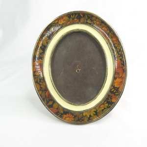May include: Oval picture frame with a floral design. The frame has a black background with orange, yellow, and brown flowers and leaves. The inner frame is cream-colored, and the photo area is brown.